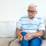 Older adult holding a painful knee while seated, representing osteoarthritis pain that may be treated with stem cell therapy.